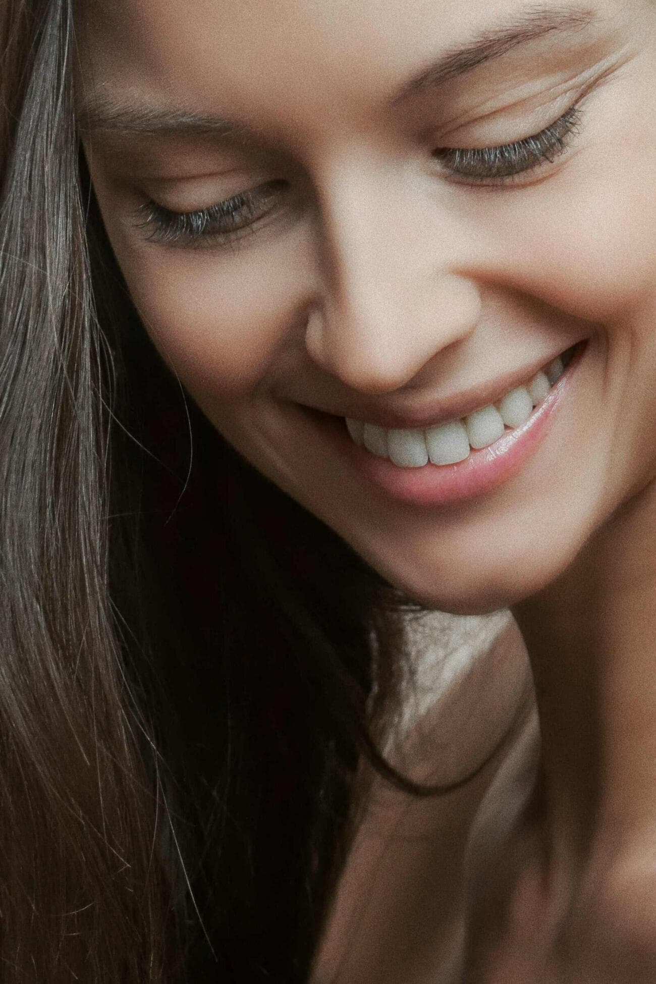A woman with long dark hair looking down and smiling, lit by soft window light