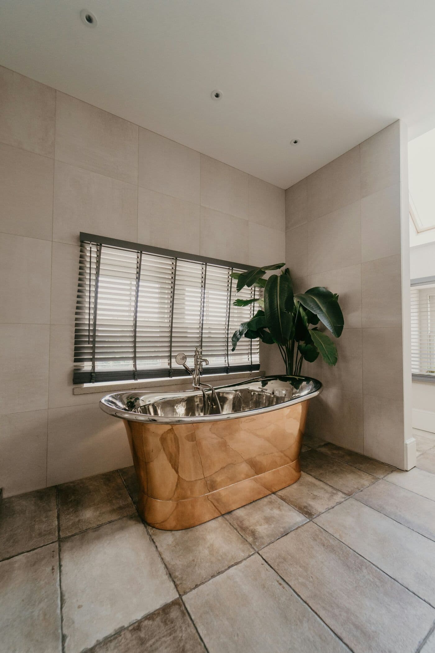 A polished copper bath beneath a shuttered window, with a tall plant beside it