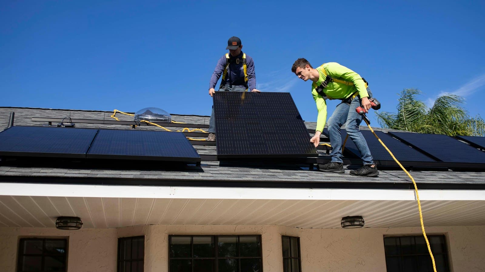 Two installers in harnesses lowering an all-black solar panel onto a roof