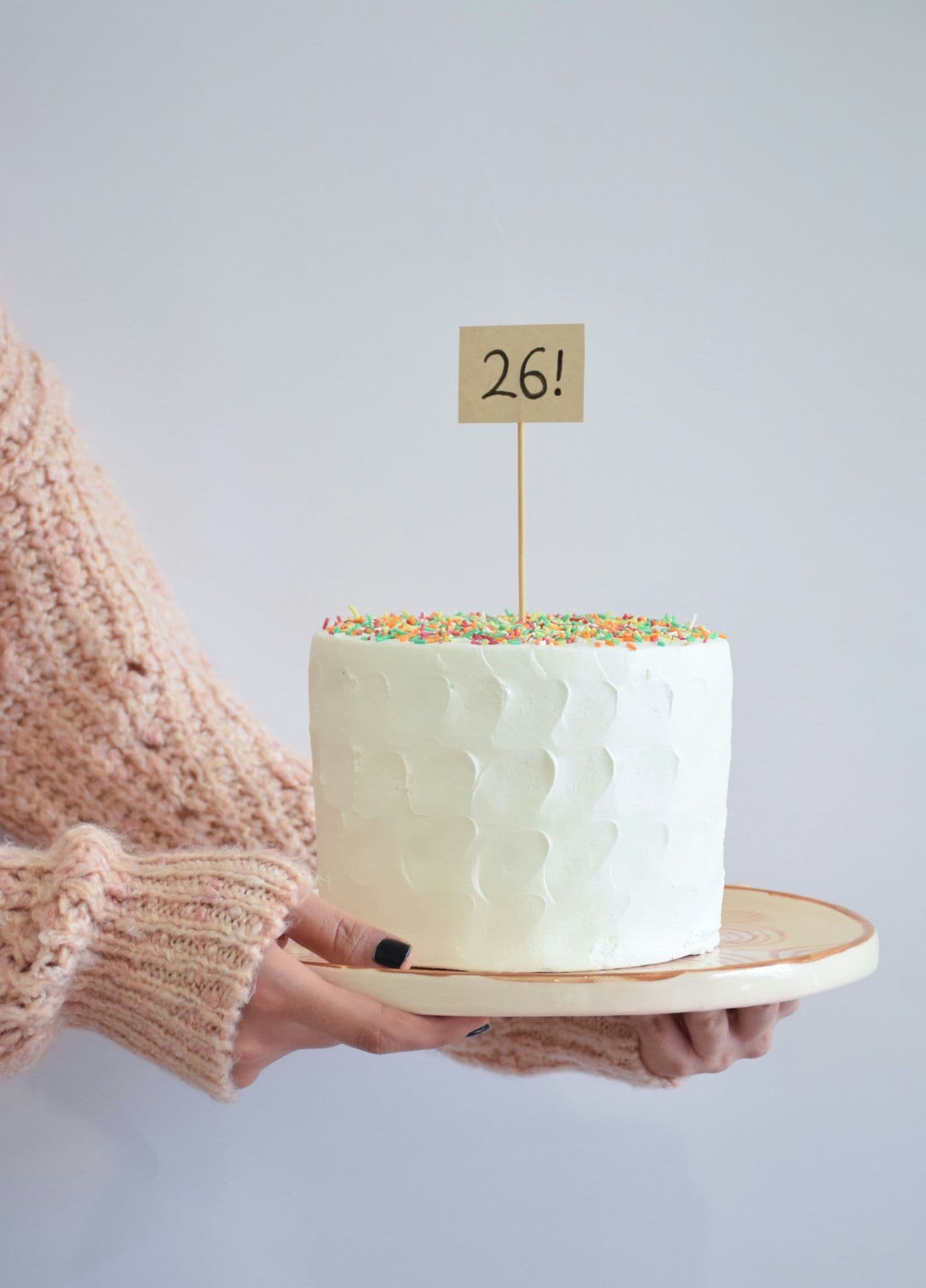 Hands holding a tall white celebration cake with sprinkles and a small number topper