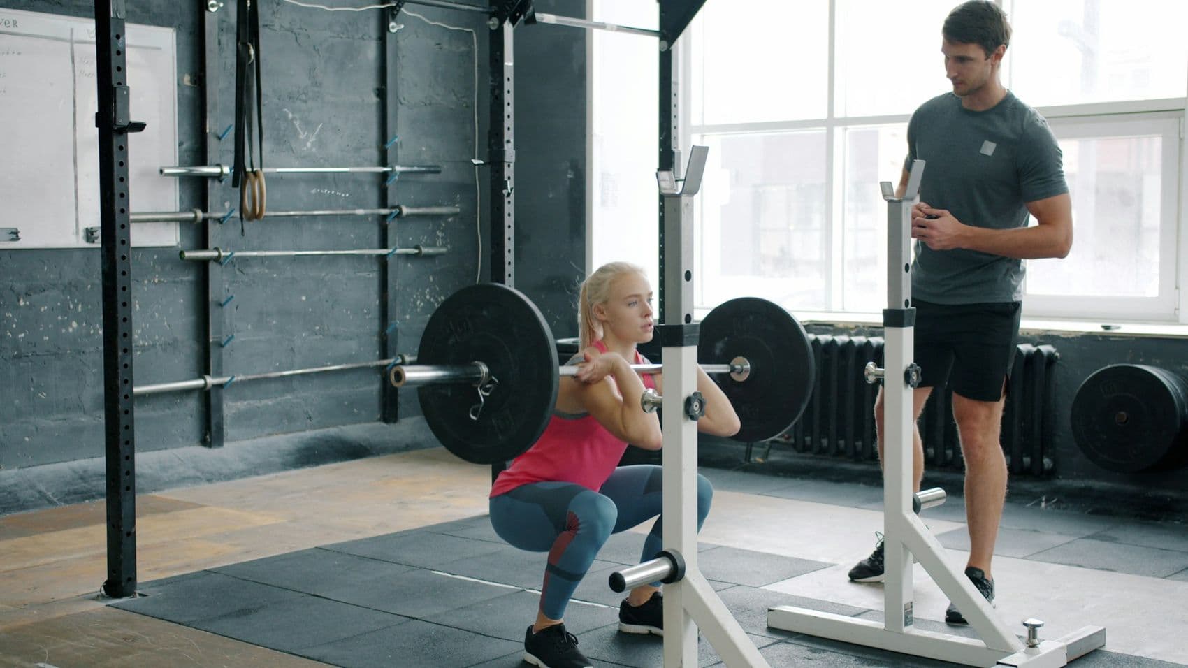 A coach watching a member squat in a bright gym with tall windows