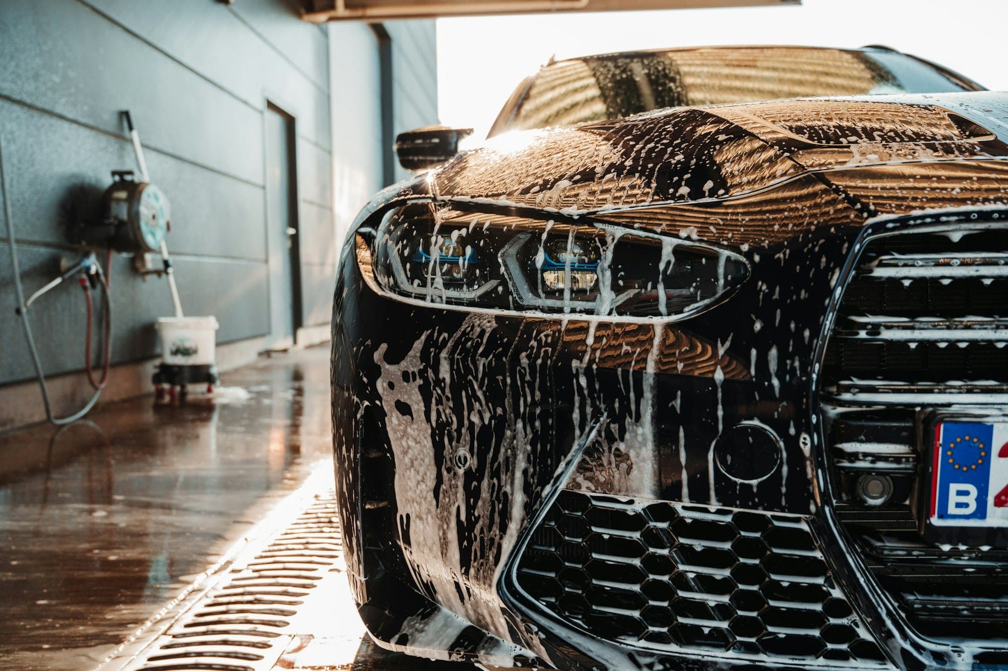 The front of a dark car streaked with foam, water running across a wet workshop floor