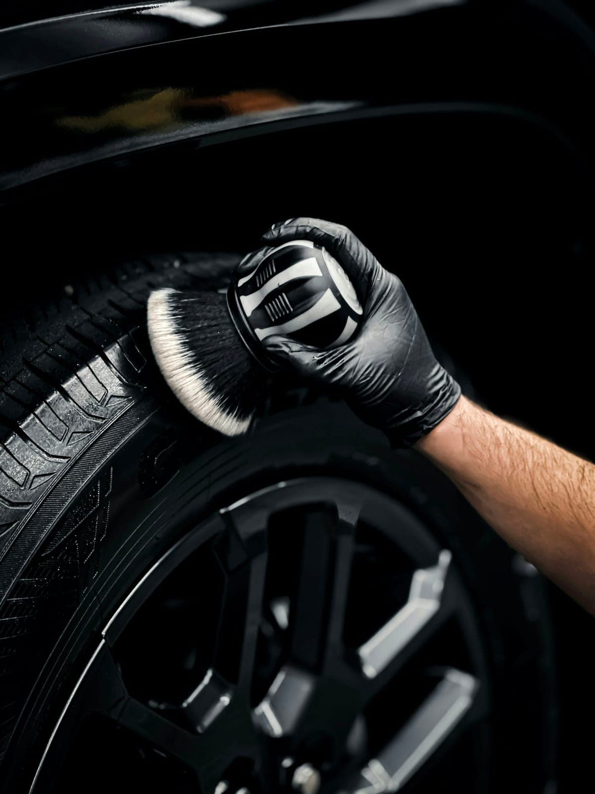 A gloved hand cleaning the spokes of a black alloy wheel with a soft brush