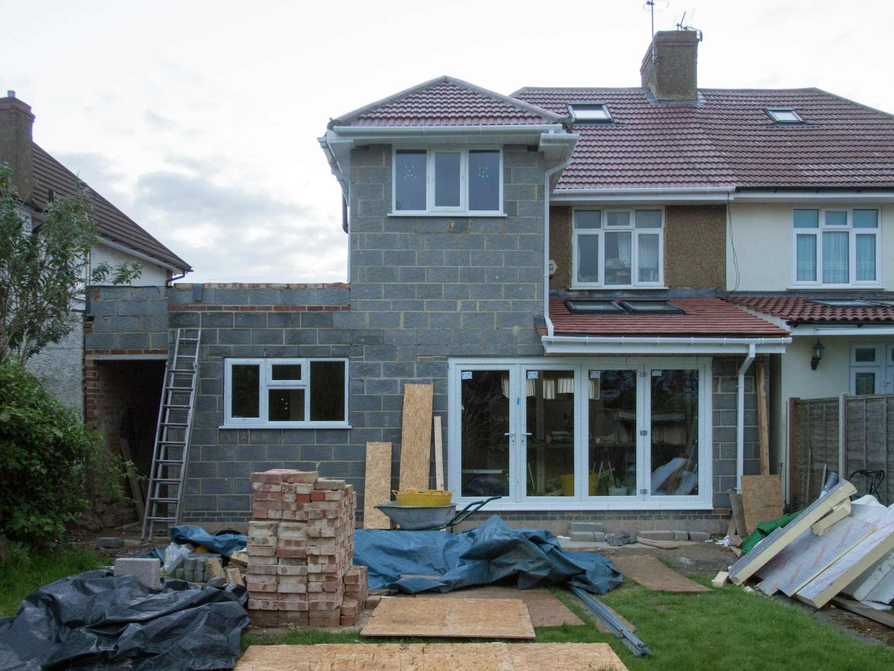 The semi-detached house mid-build: blockwork shell of the new rear extension, scaffold boards and sandbags on the lawn