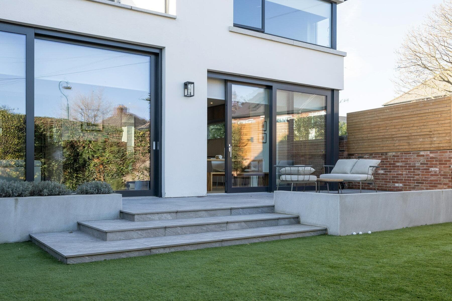 A white rendered two storey rear extension with large dark-framed sliding doors and stone steps down to a lawn