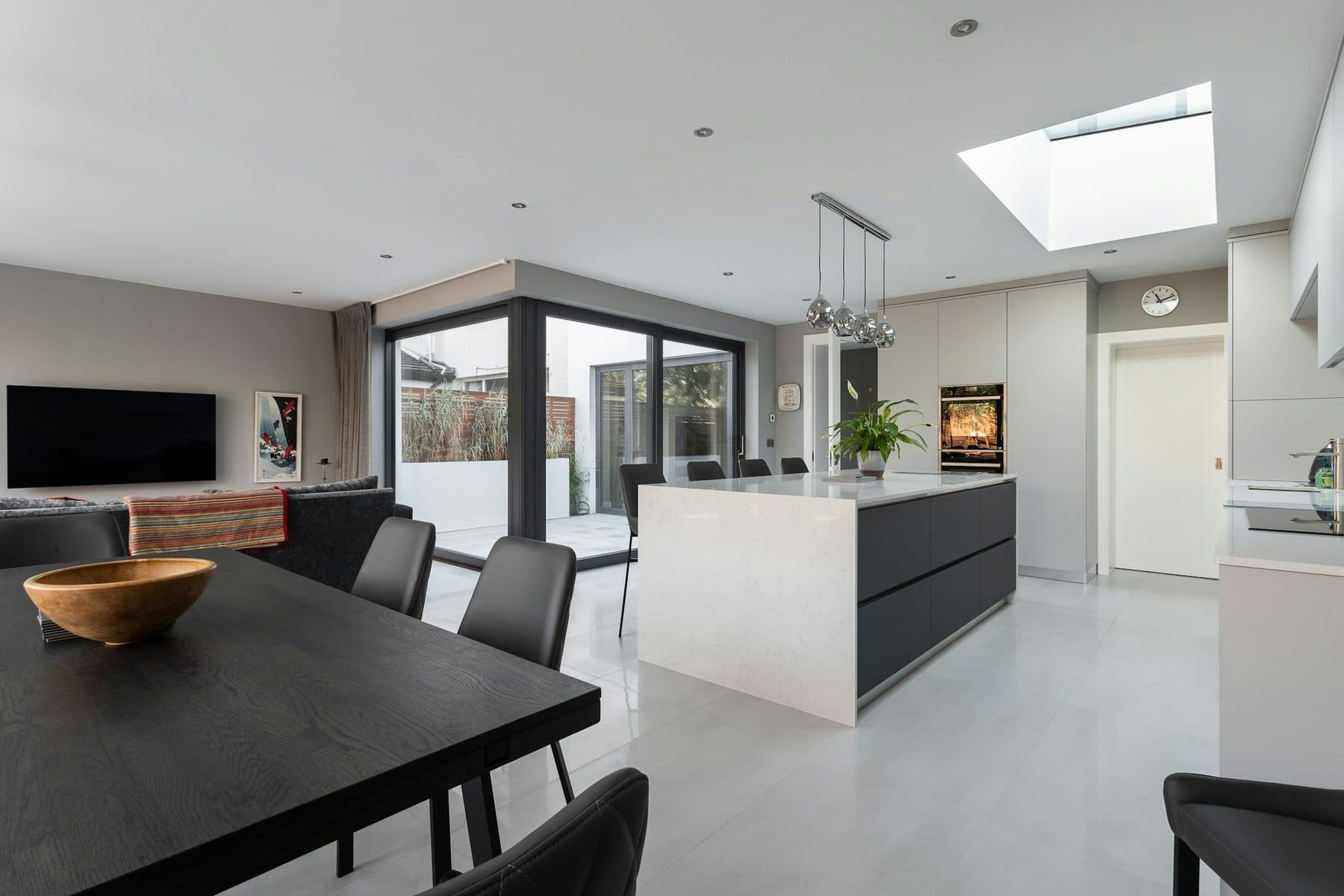 An open-plan kitchen with a dark island, a dining table and floor-to-ceiling glazing to the garden