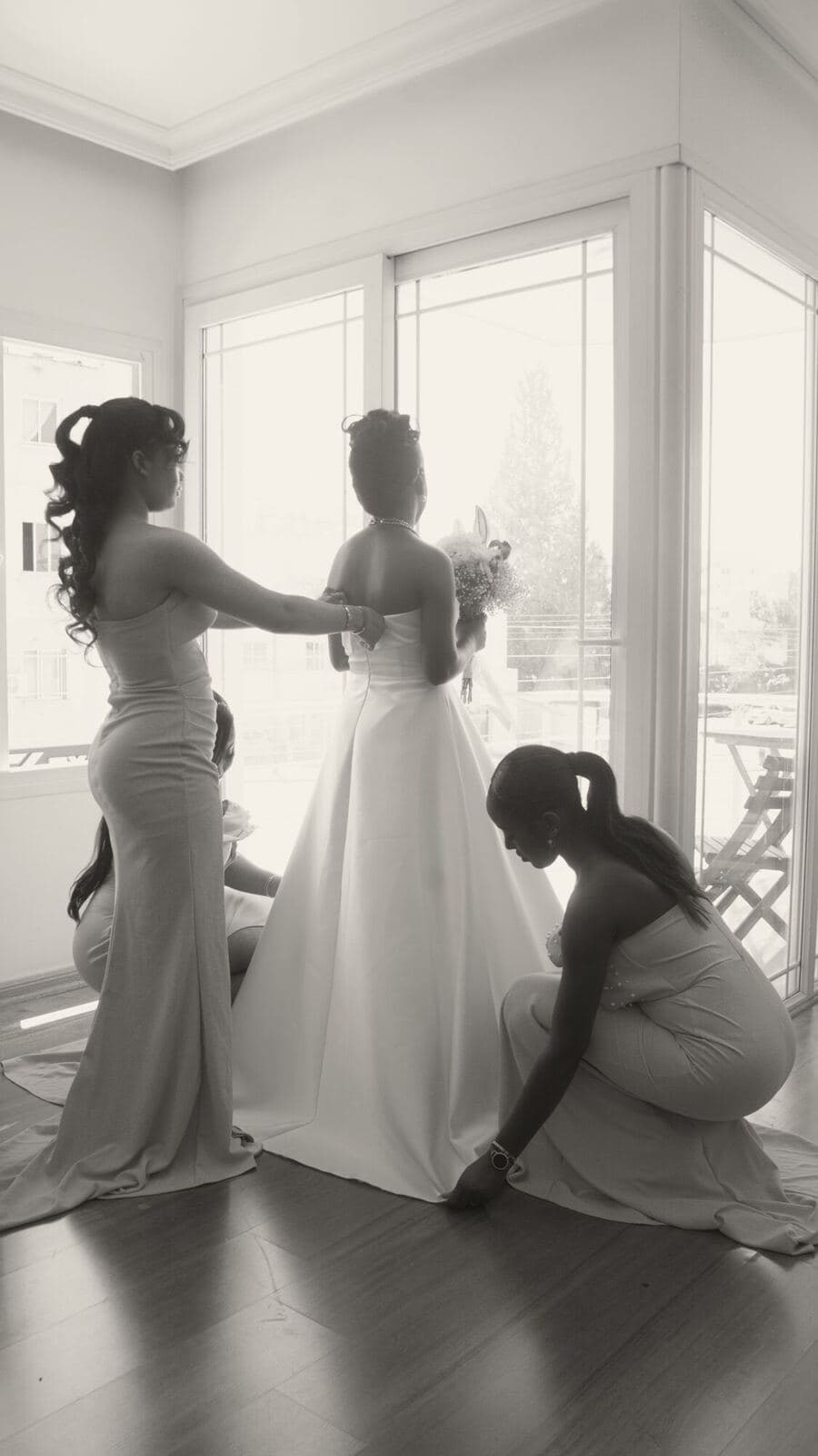 Bridesmaids fasten the bride’s dress by a bright window