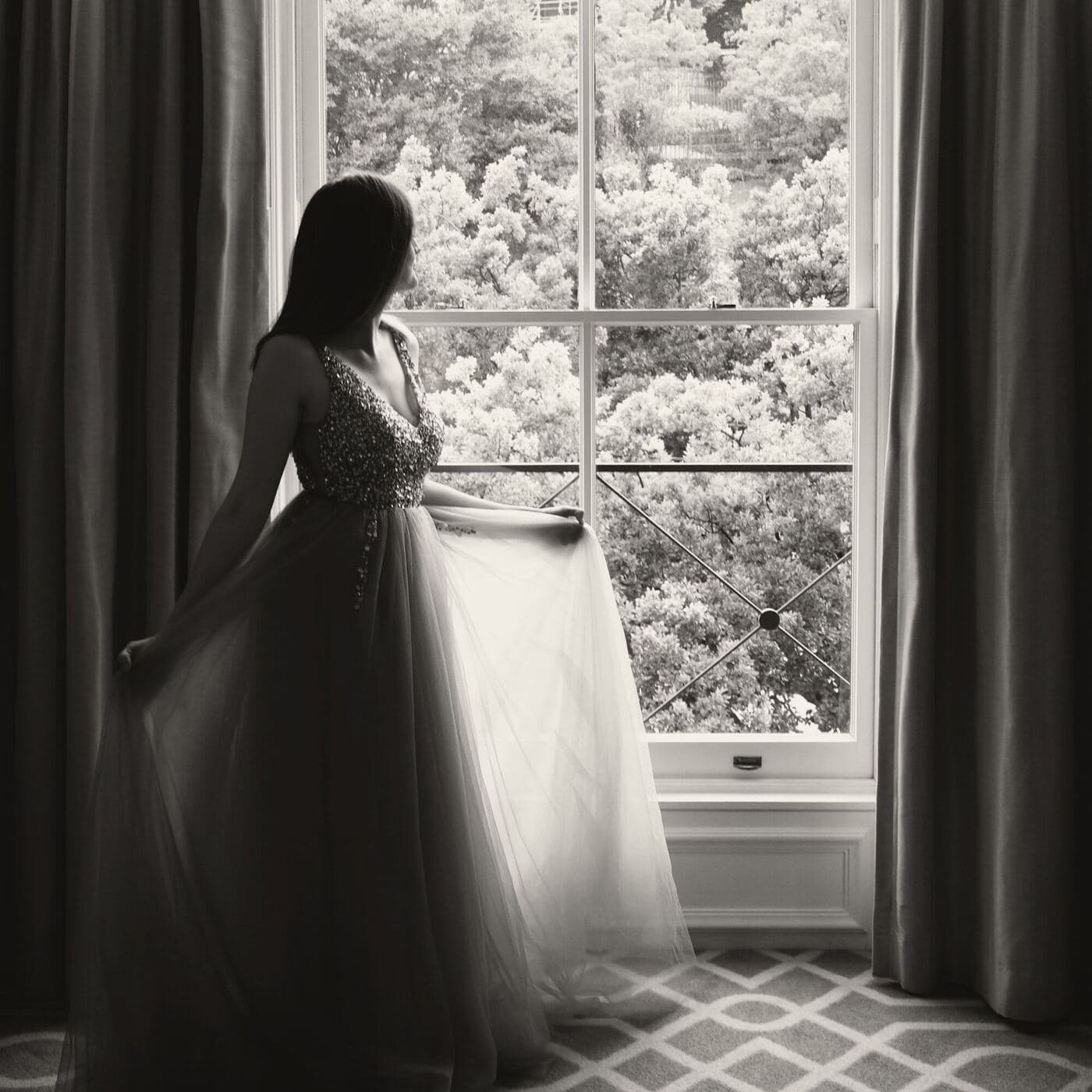 A bride in her gown stands at a tall sash window, the garden bright behind her