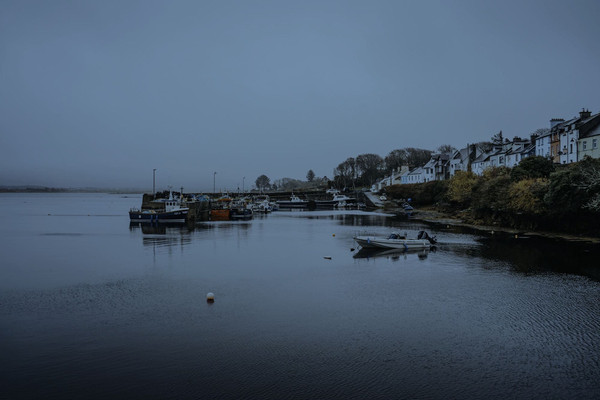 A row of whitewashed cottages above a still harbour at blue hour, fishing boats moored under a misty sky