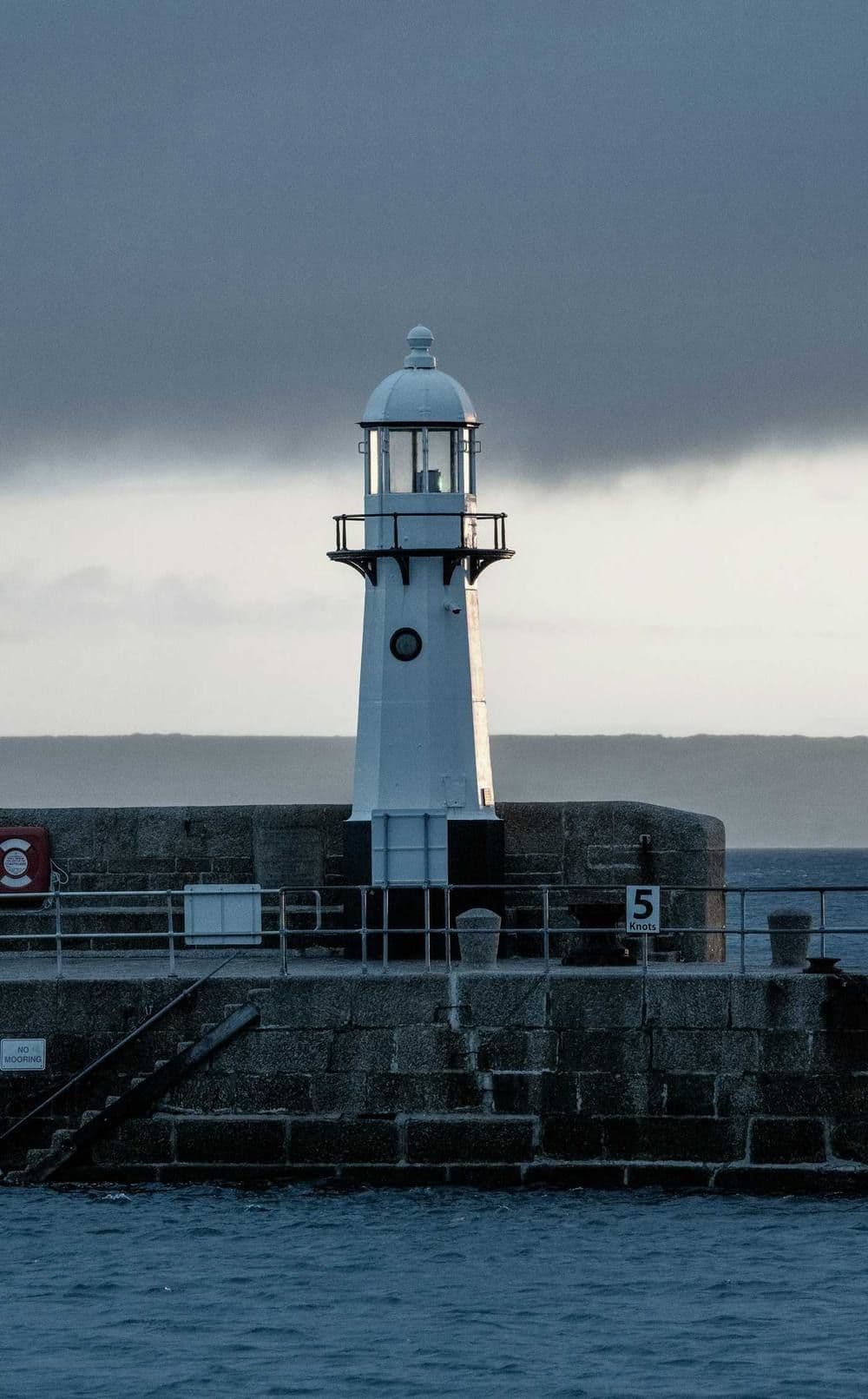 A small white lighthouse at the end of a granite pier under a grey sky