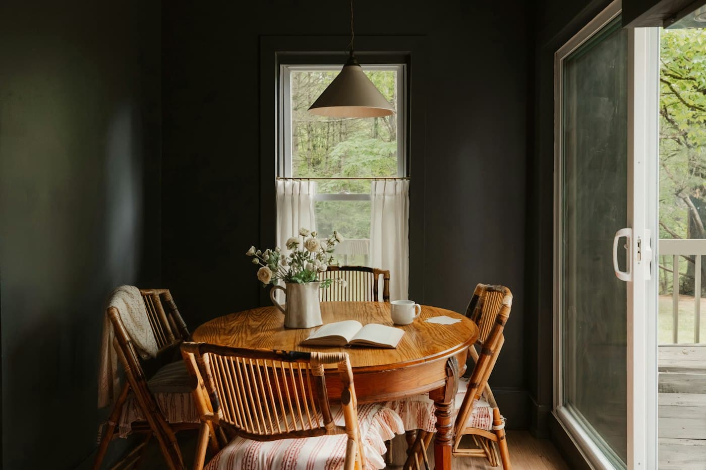 A round table and cane chairs in a dark painted dining room beside a sunlit window