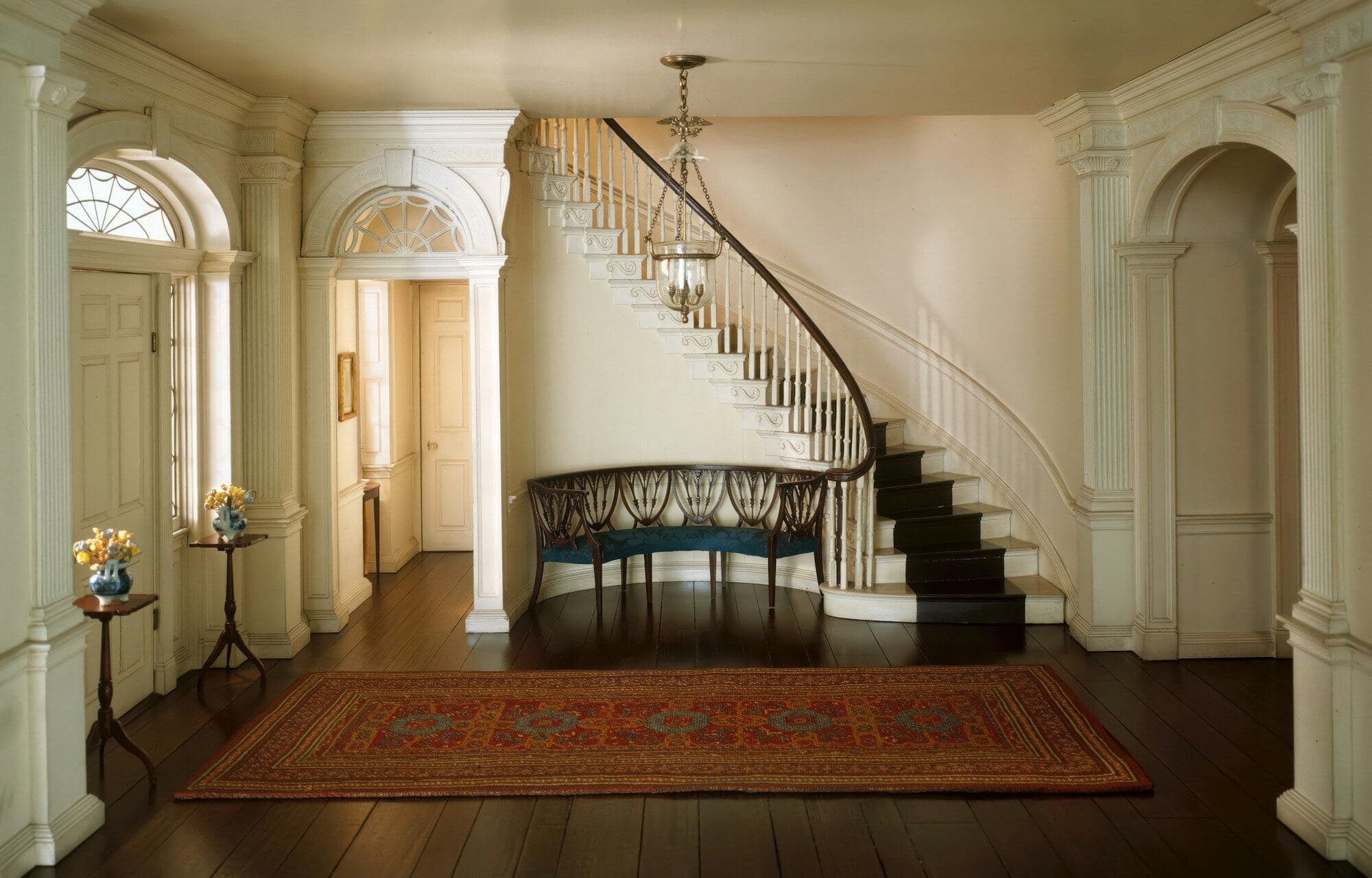 A cream Georgian entrance hall with arched doorways, a curving staircase and a rug on dark boards