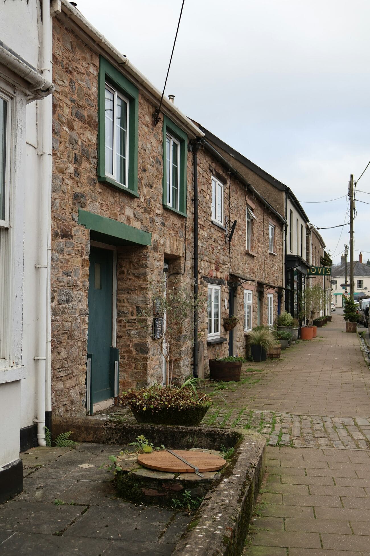 A quiet village pavement lined with stone cottages, green window frames and pots by the doors
