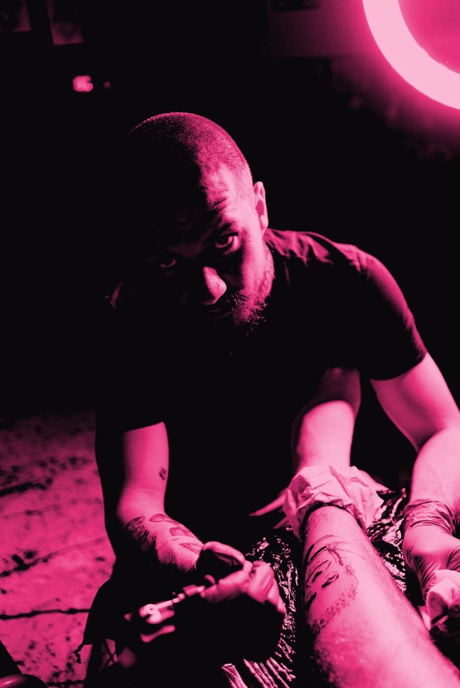 A tattooist with a shaved head leaning over a client's arm, working under a ring light