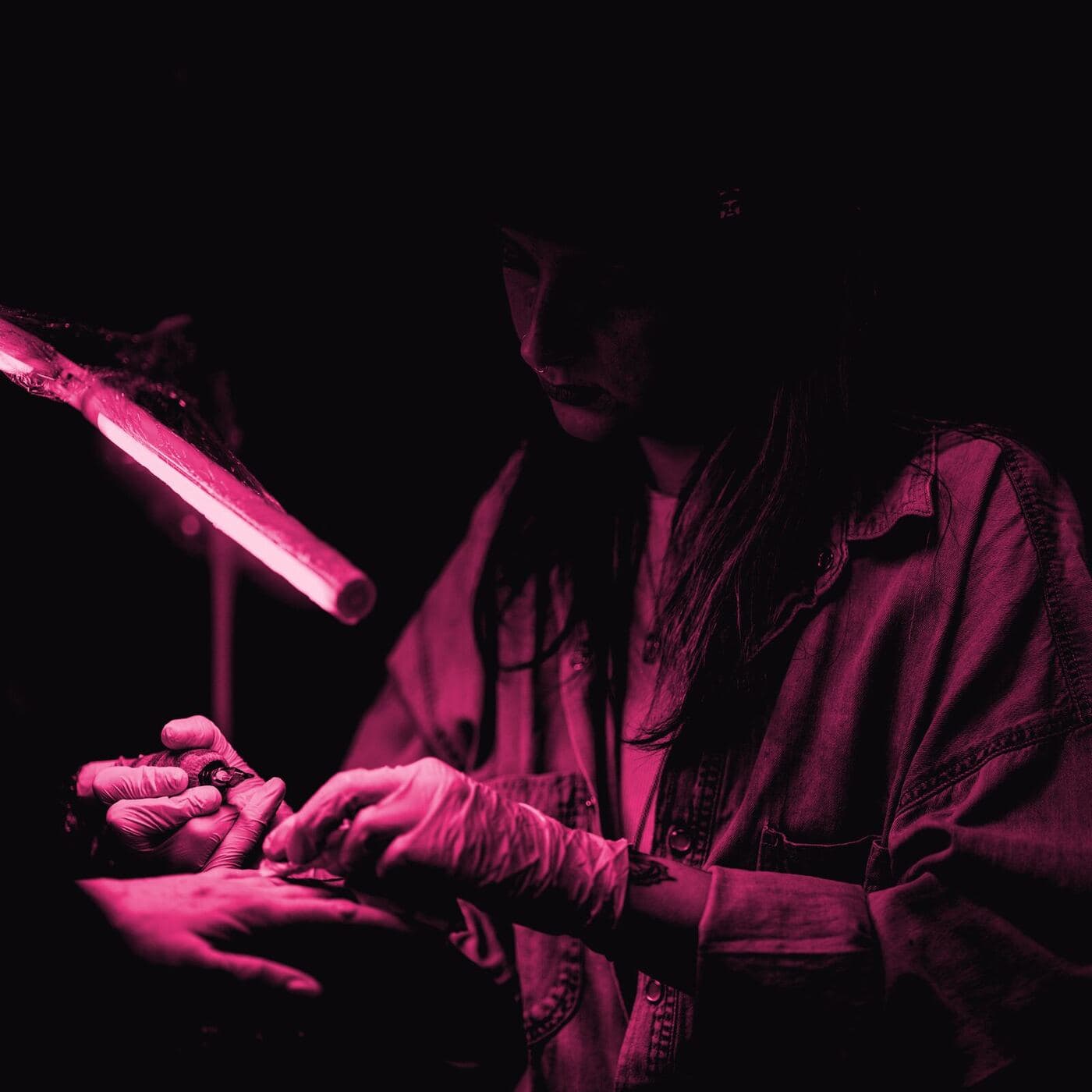 A tattooist in gloves working on a small design on a client's hand under a strip light