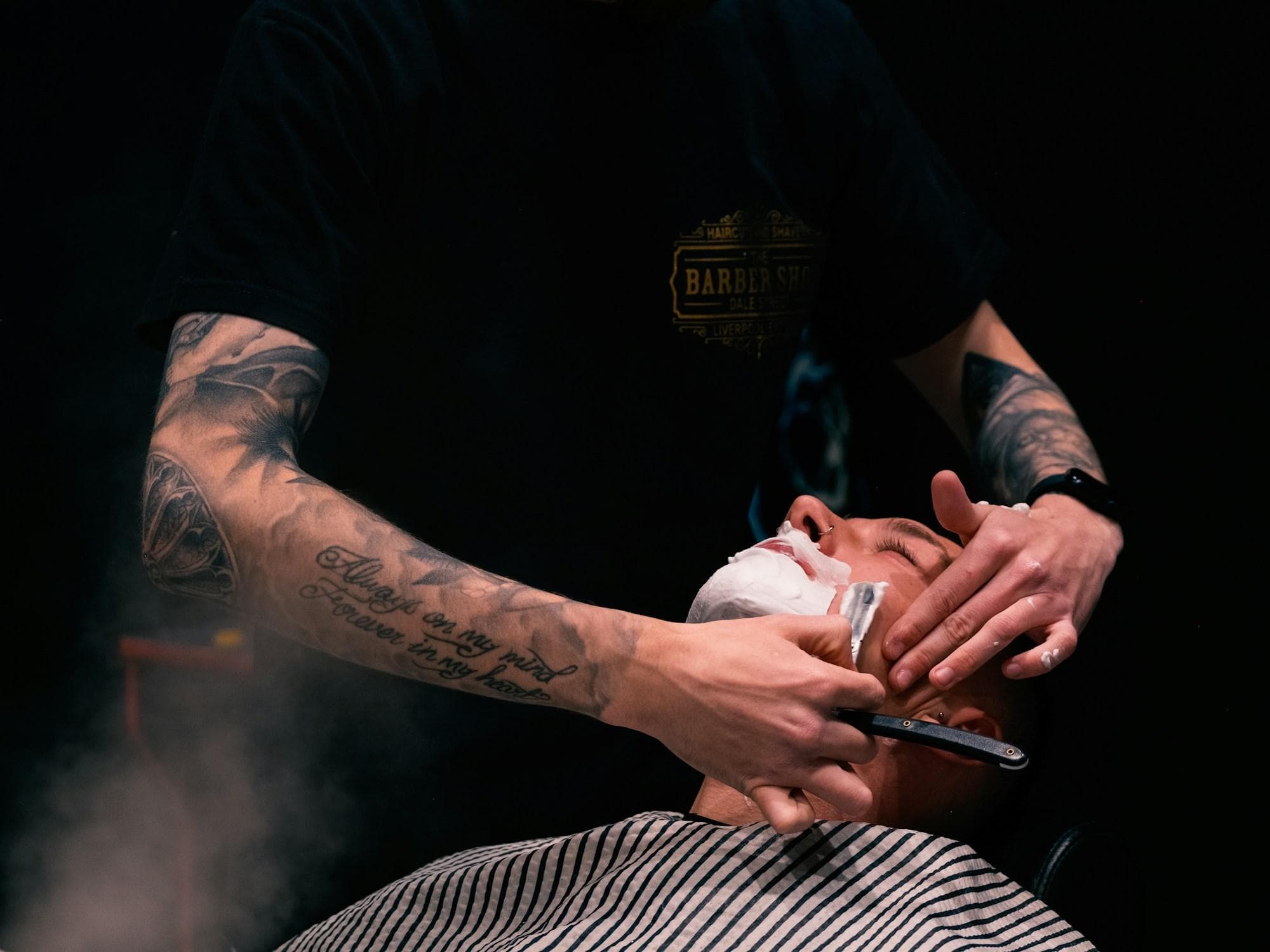 A tattooed barber giving a cut-throat razor shave through hot lather, steam rising