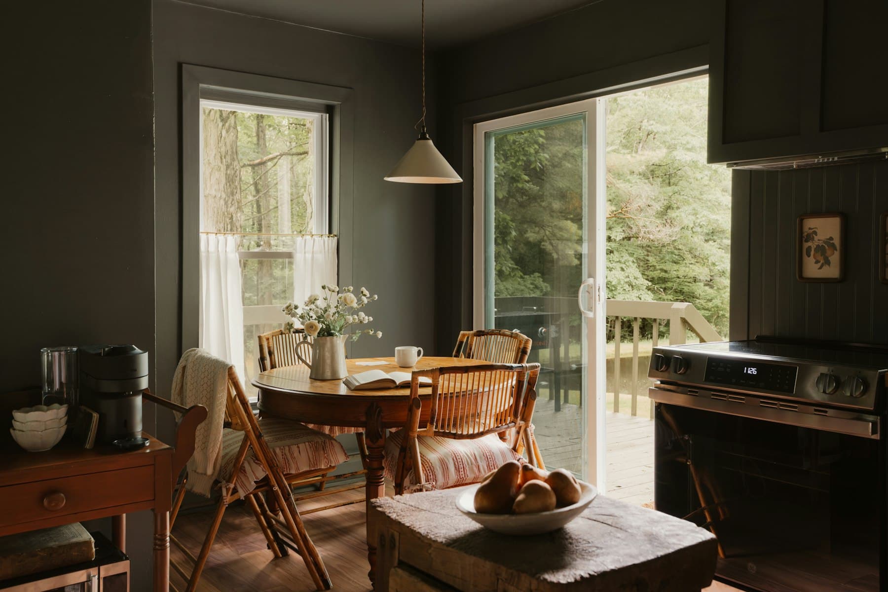 A round wooden table set by a window in a dark painted kitchen, with flowers and a bowl of bread