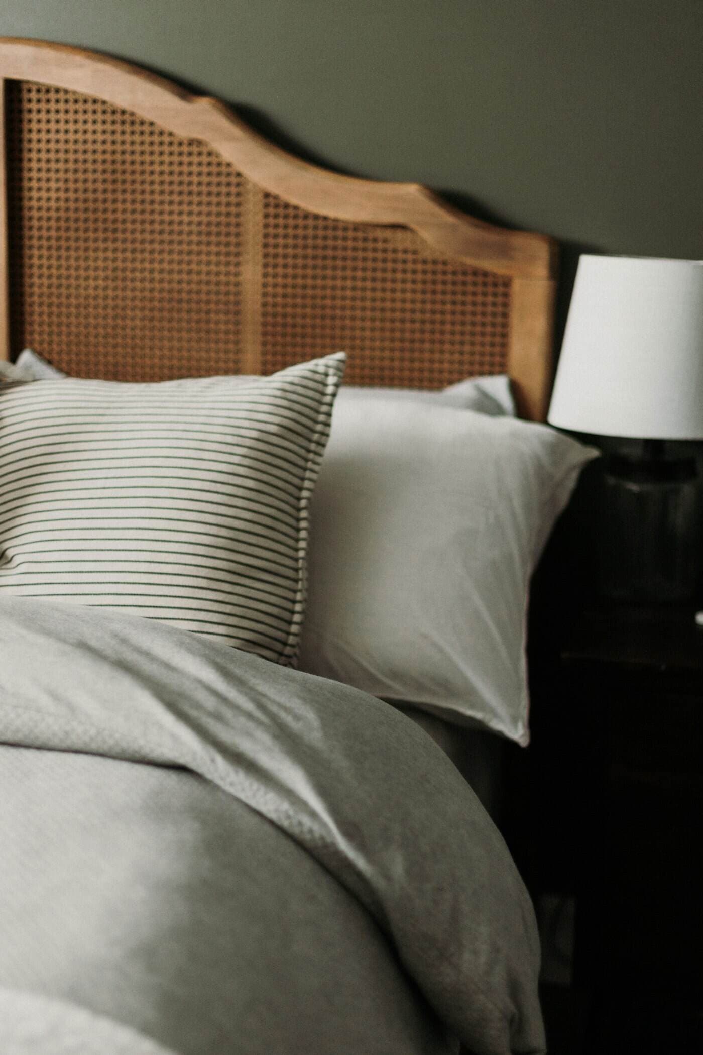 A bed with a caned walnut headboard, striped and grey linen pillows and a white bedside lamp