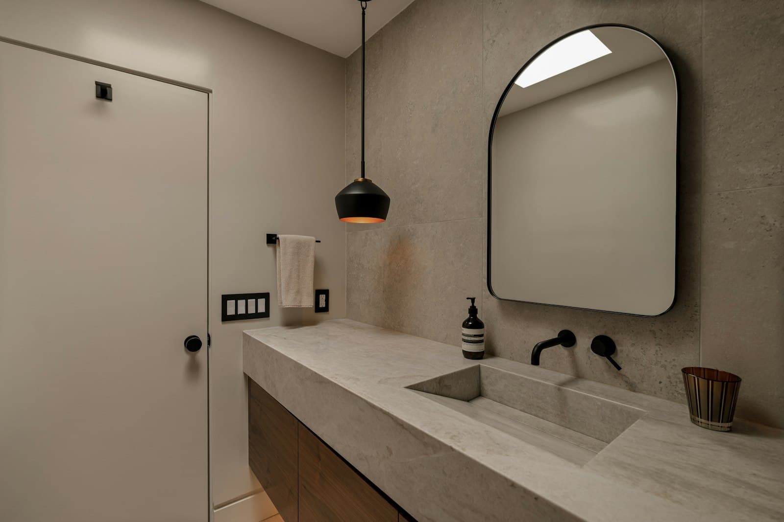 A calm stone bathroom with a long honed basin, black wall taps, an arched mirror and a black pendant light