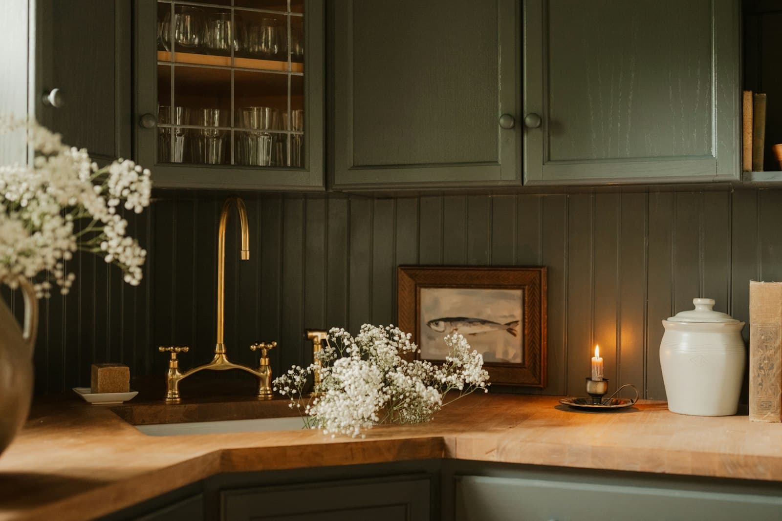 A kitchen with deep green painted cabinets, a brass bridge tap, an oak worktop and white flowers in a jug