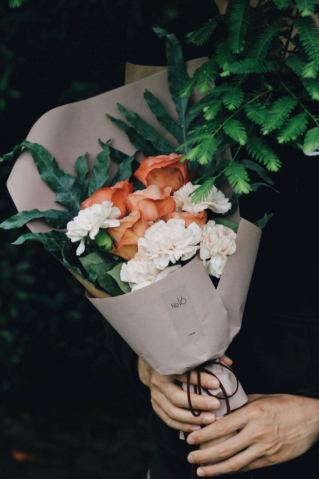 A hand-tied bouquet of peach roses and white carnations wrapped in pale paper, held in both hands