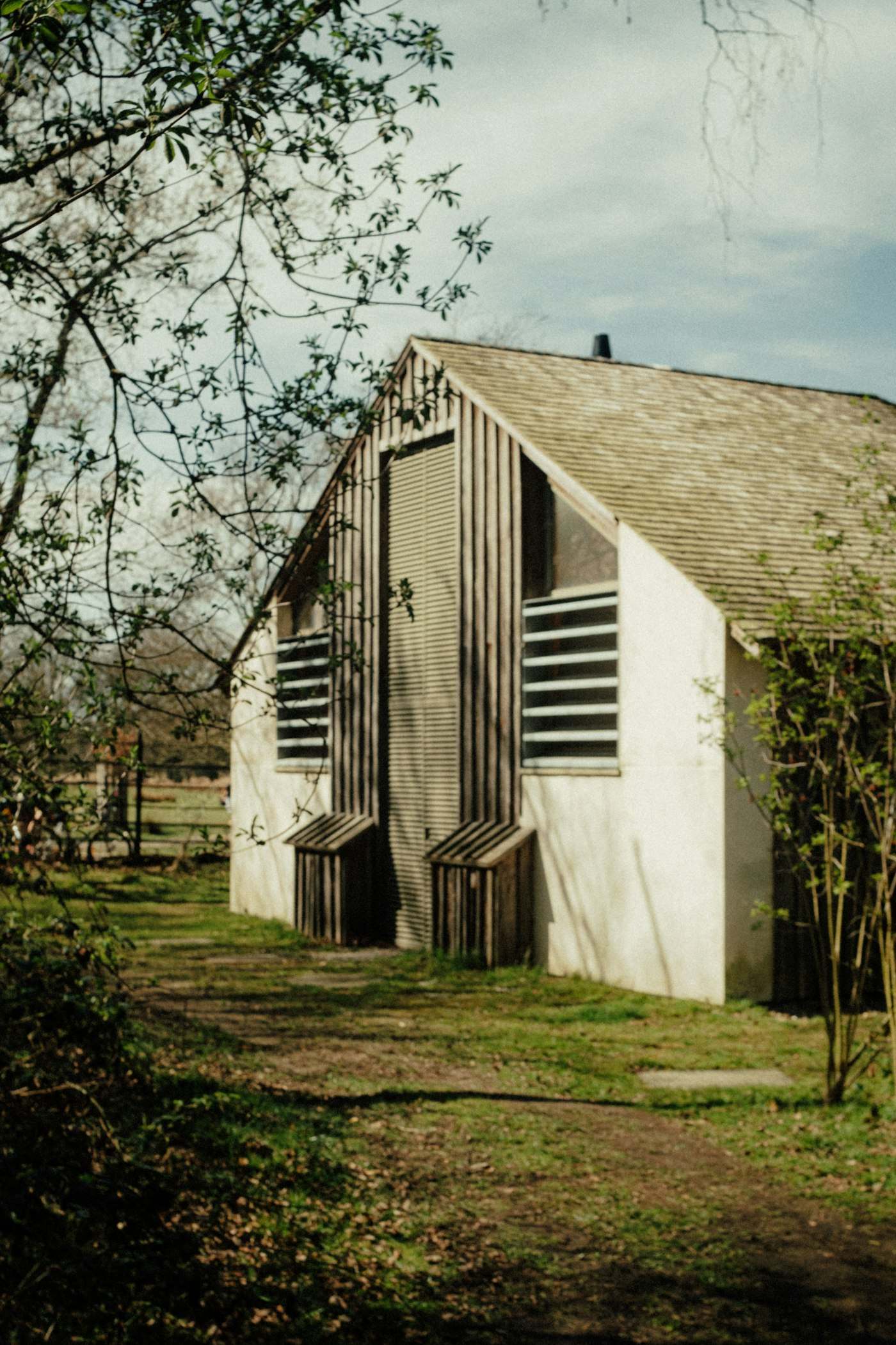 A pale timber barn with a steep gable and slatted openings, among bare winter trees