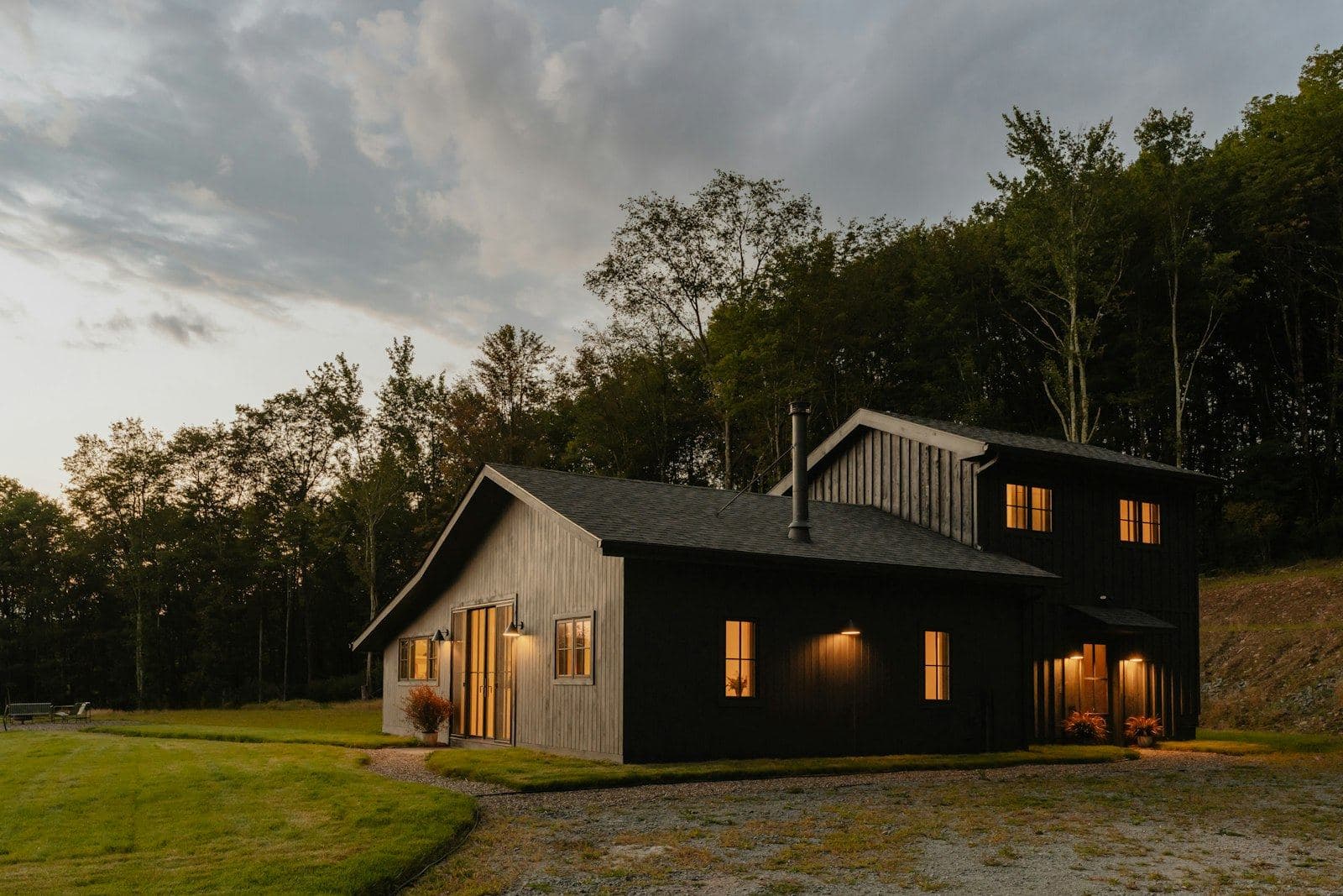 A dark-clad house with warm windows sitting low in a meadow below woodland