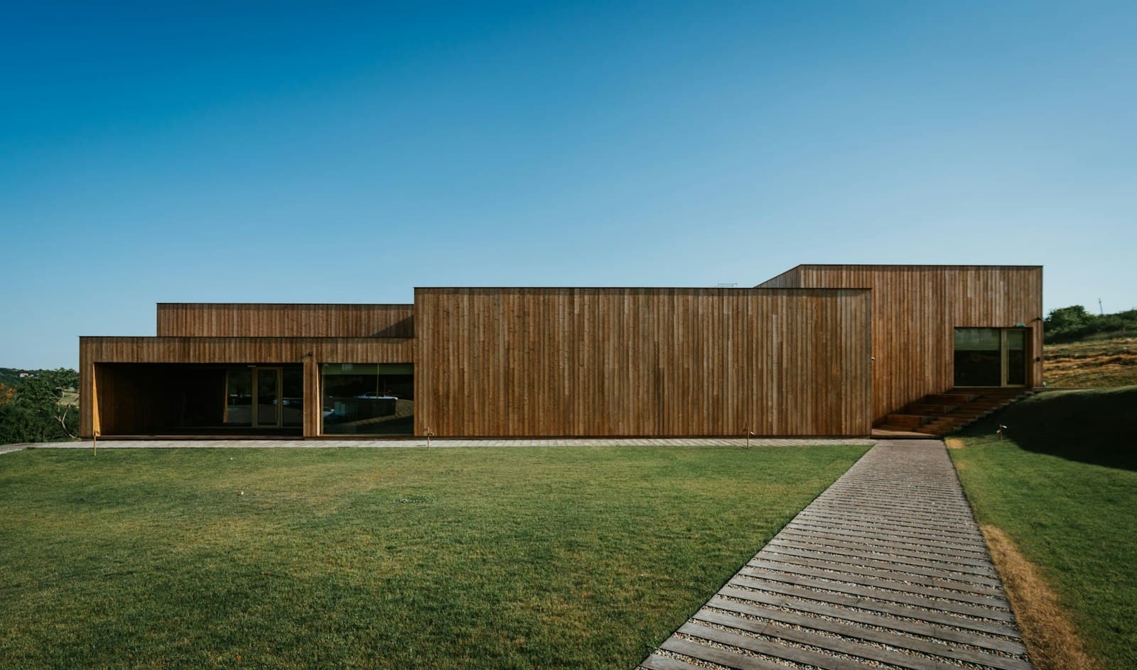 A long, low timber-clad house with a flat roof and a boardwalk across the lawn to the entrance