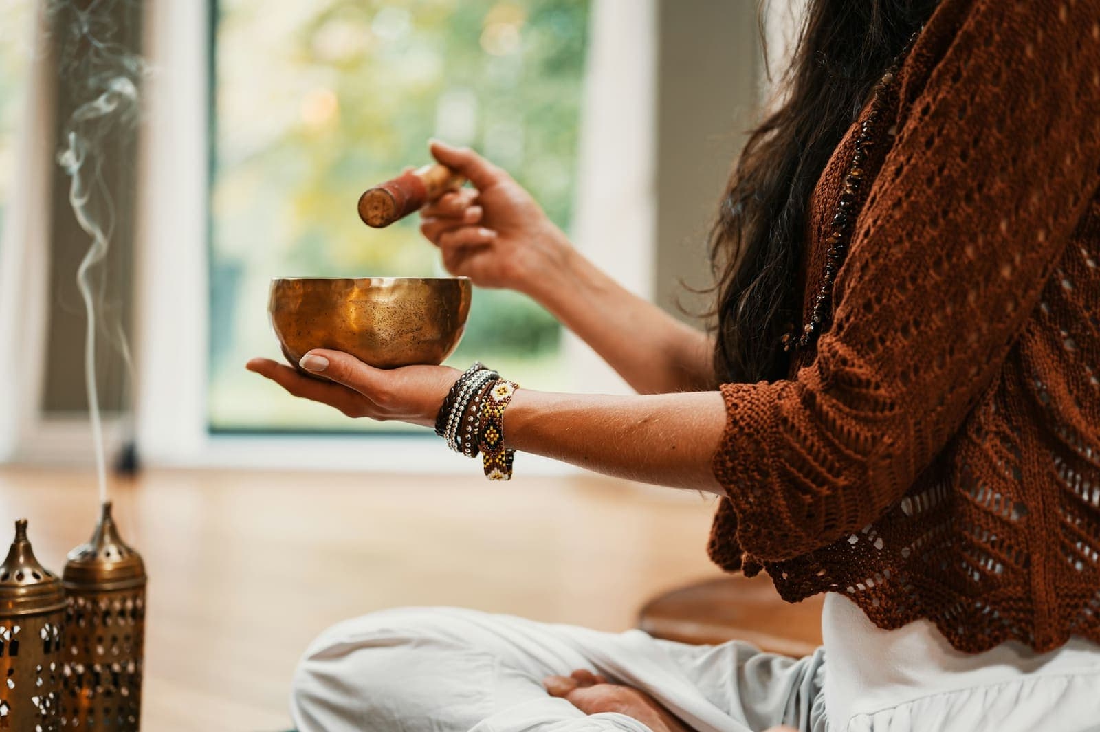 A person sitting cross-legged holding a brass singing bowl, incense beside them
