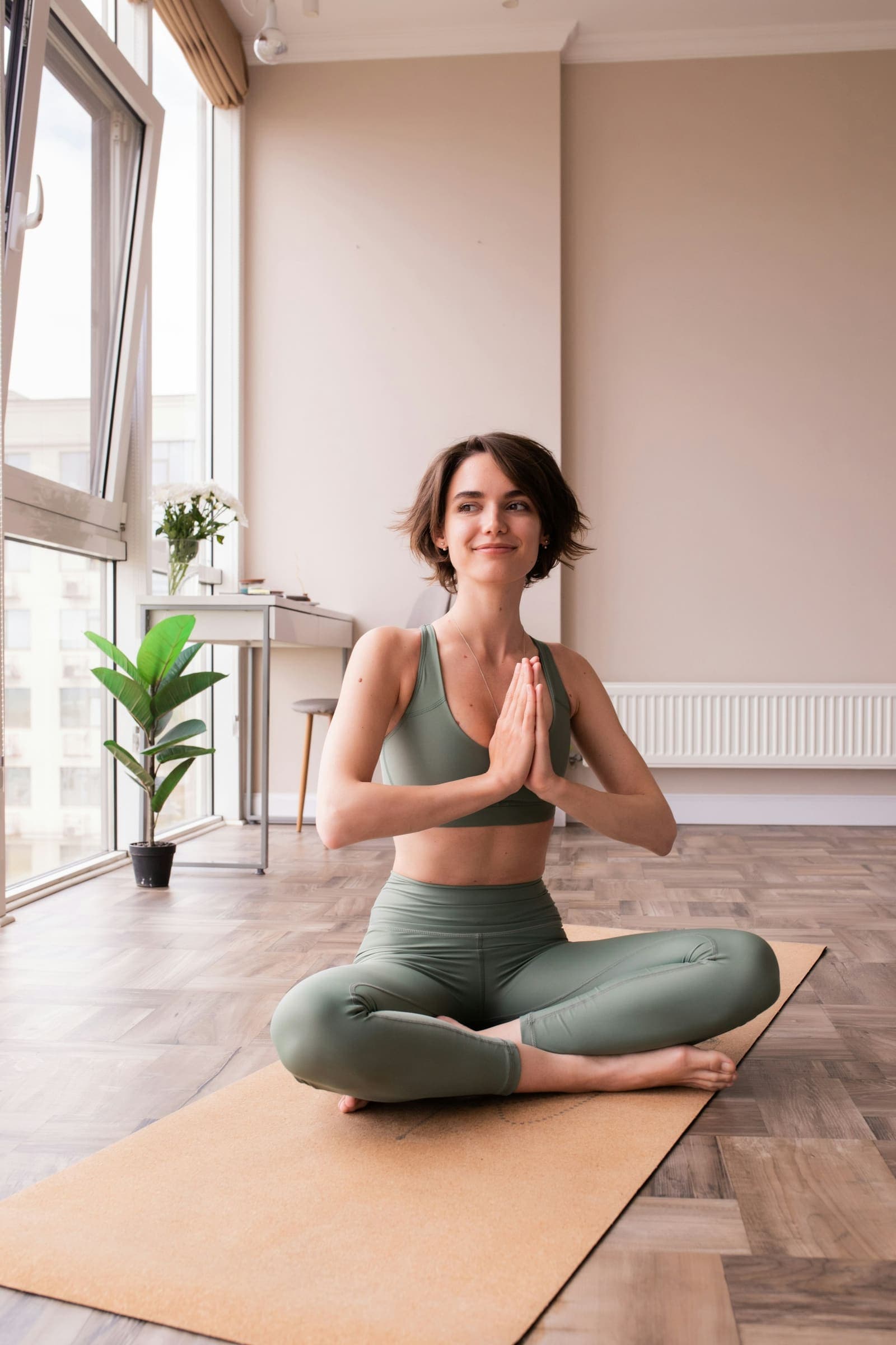 A woman sitting cross-legged with her hands together in a bright room with plants