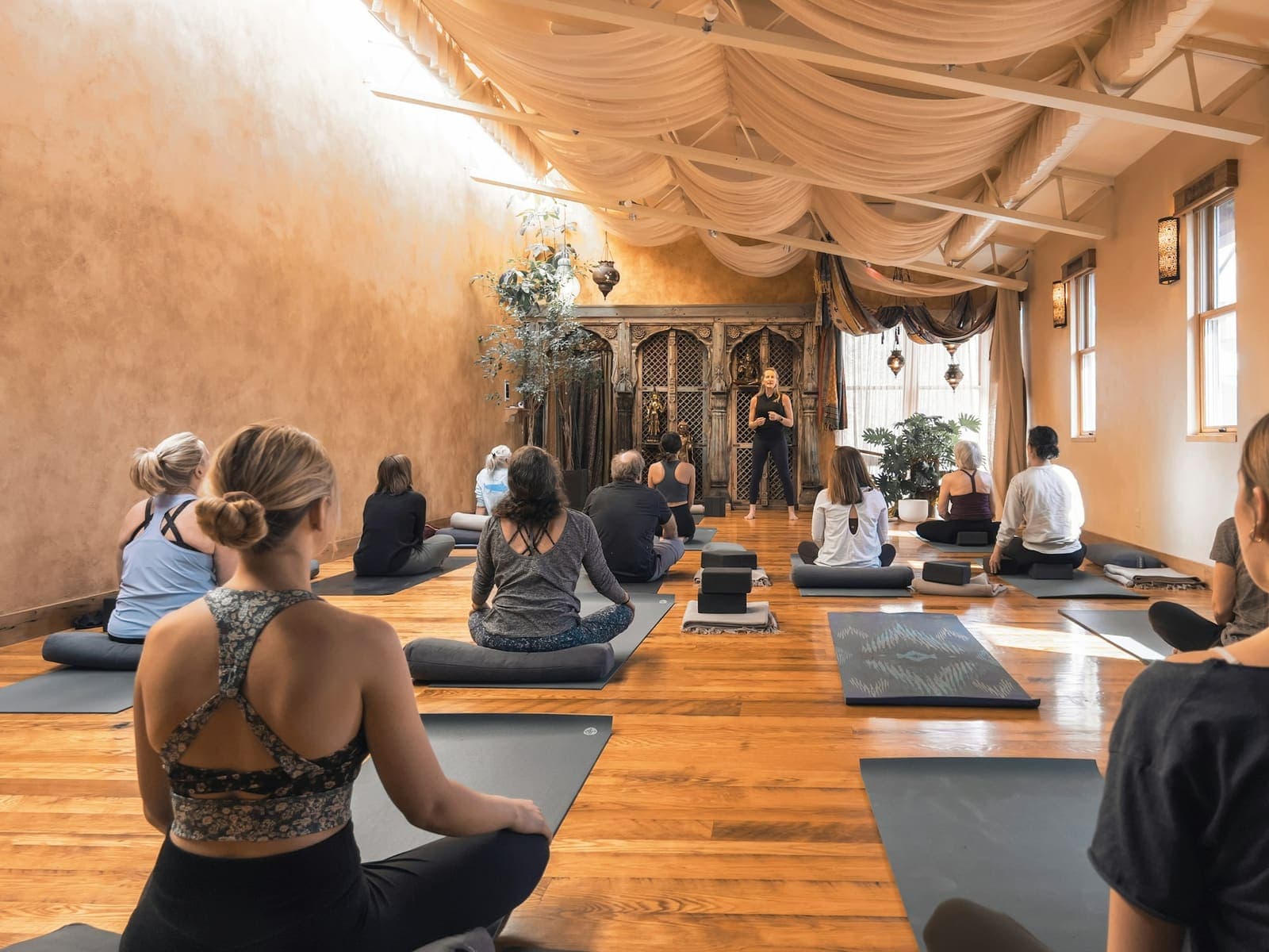 A full yoga class seated on mats in a warm, wood-floored studio with soft light