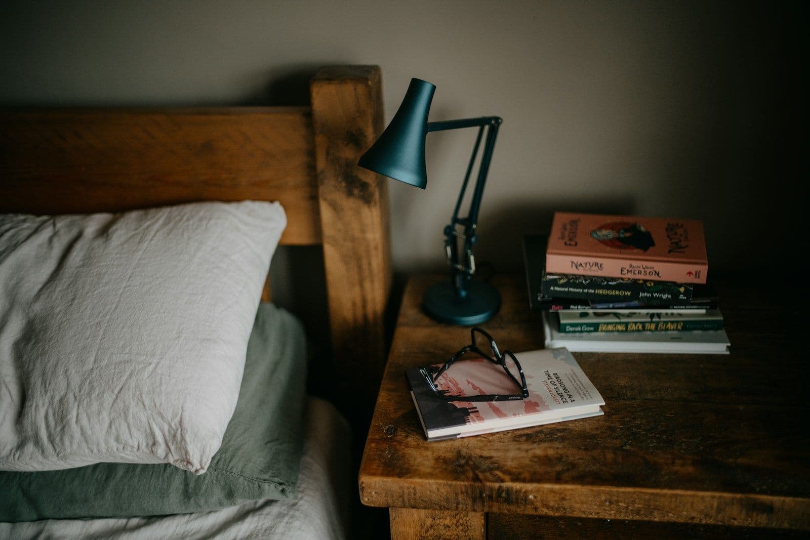 An oak bedhead and bedside table with an anglepoise lamp, a stack of books and reading glasses
