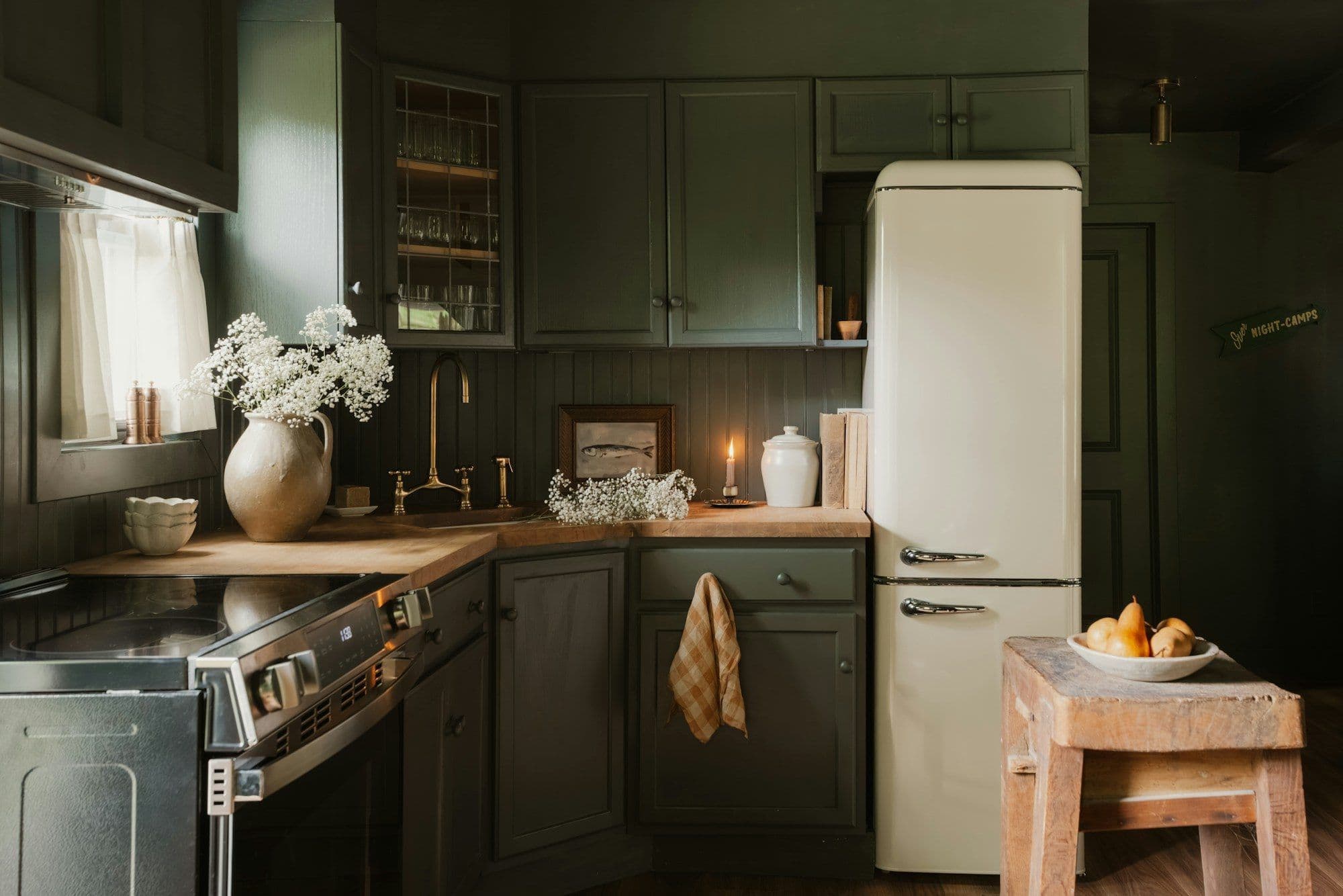 A deep green painted kitchen with an oak worktop, brass taps and a cream fridge, lit by a window